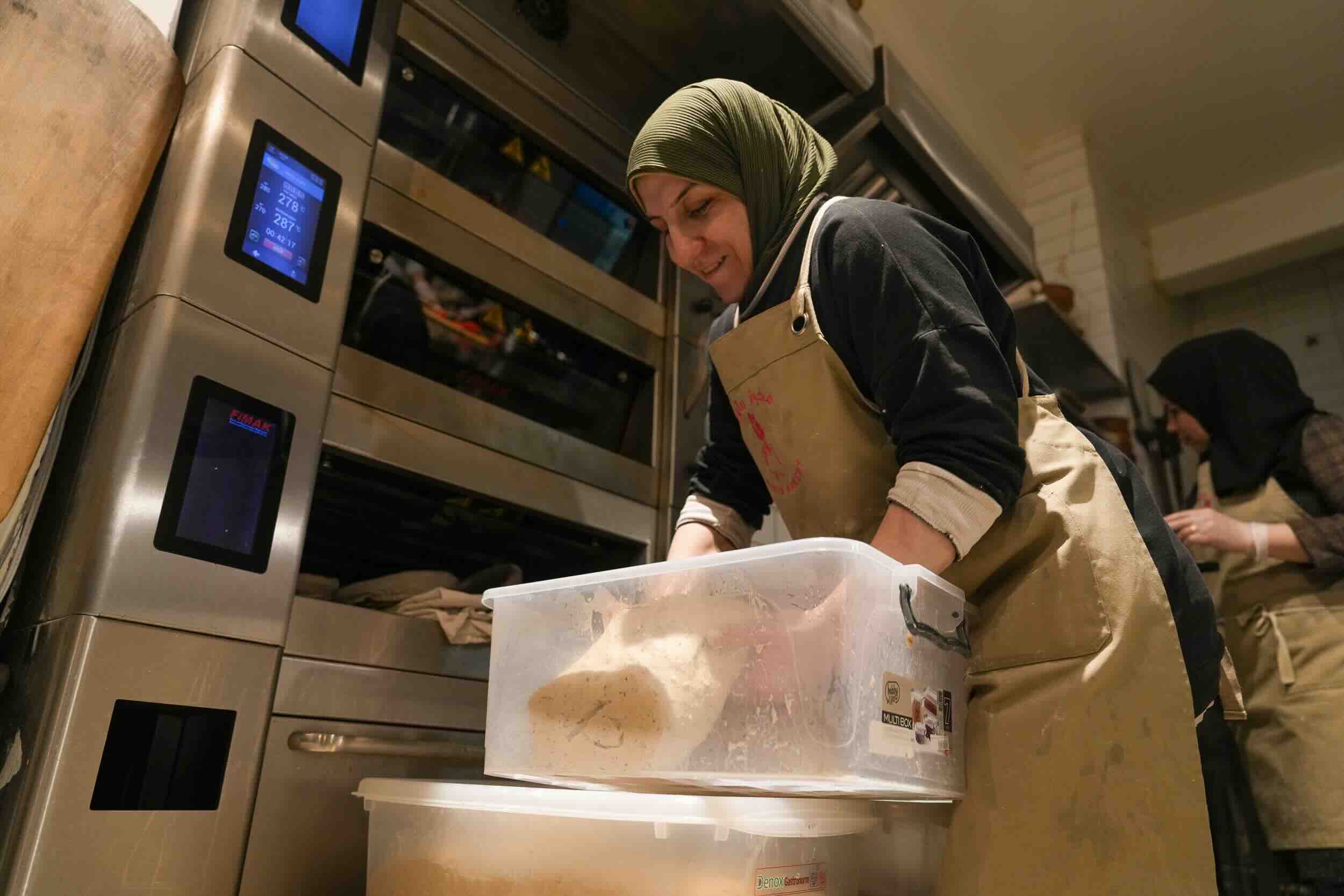 Image of a lady wearing headscarf lifting a bread dough from a box in a bakery