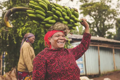 Image of a female person in the street carrying a bunch of banana on her head and smiling with another person at the background